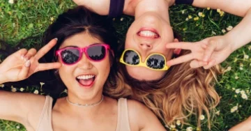 Two smiling people wearing sunglasses. They are making peace signs and lying on their backs in the grass.