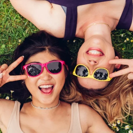 Two smiling people wearing sunglasses. They are making peace signs and lying on their backs in the grass.